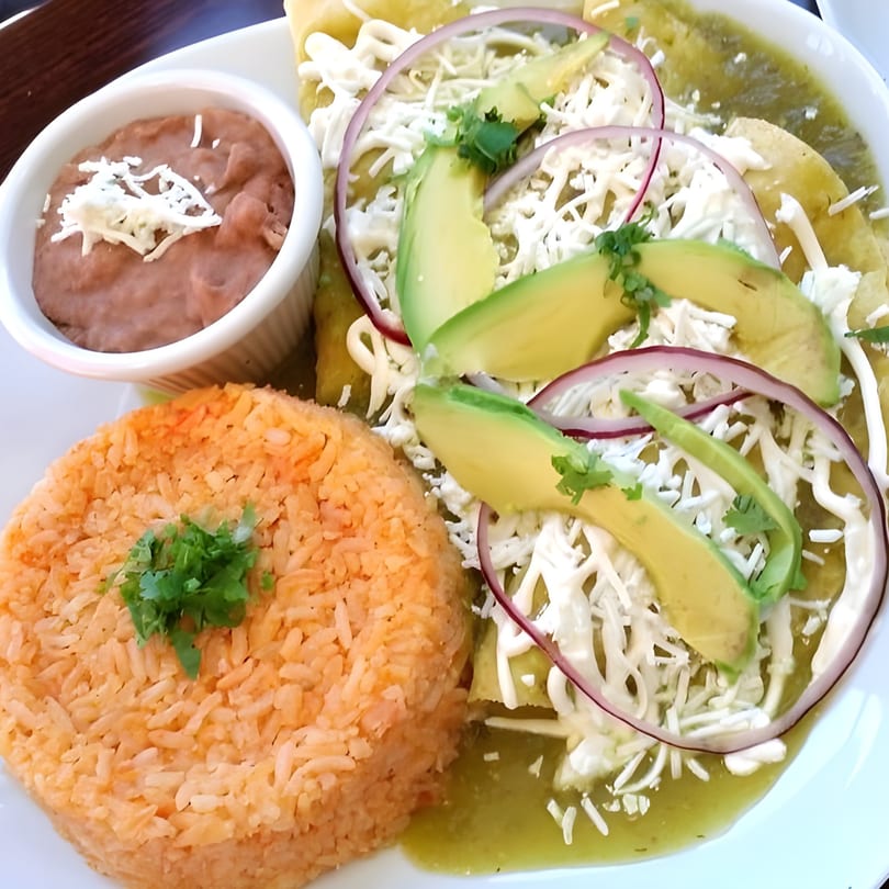 A plate of Sol Azteca's enchiladas with green sauce, rice, refried beans, avocado, and red onion.